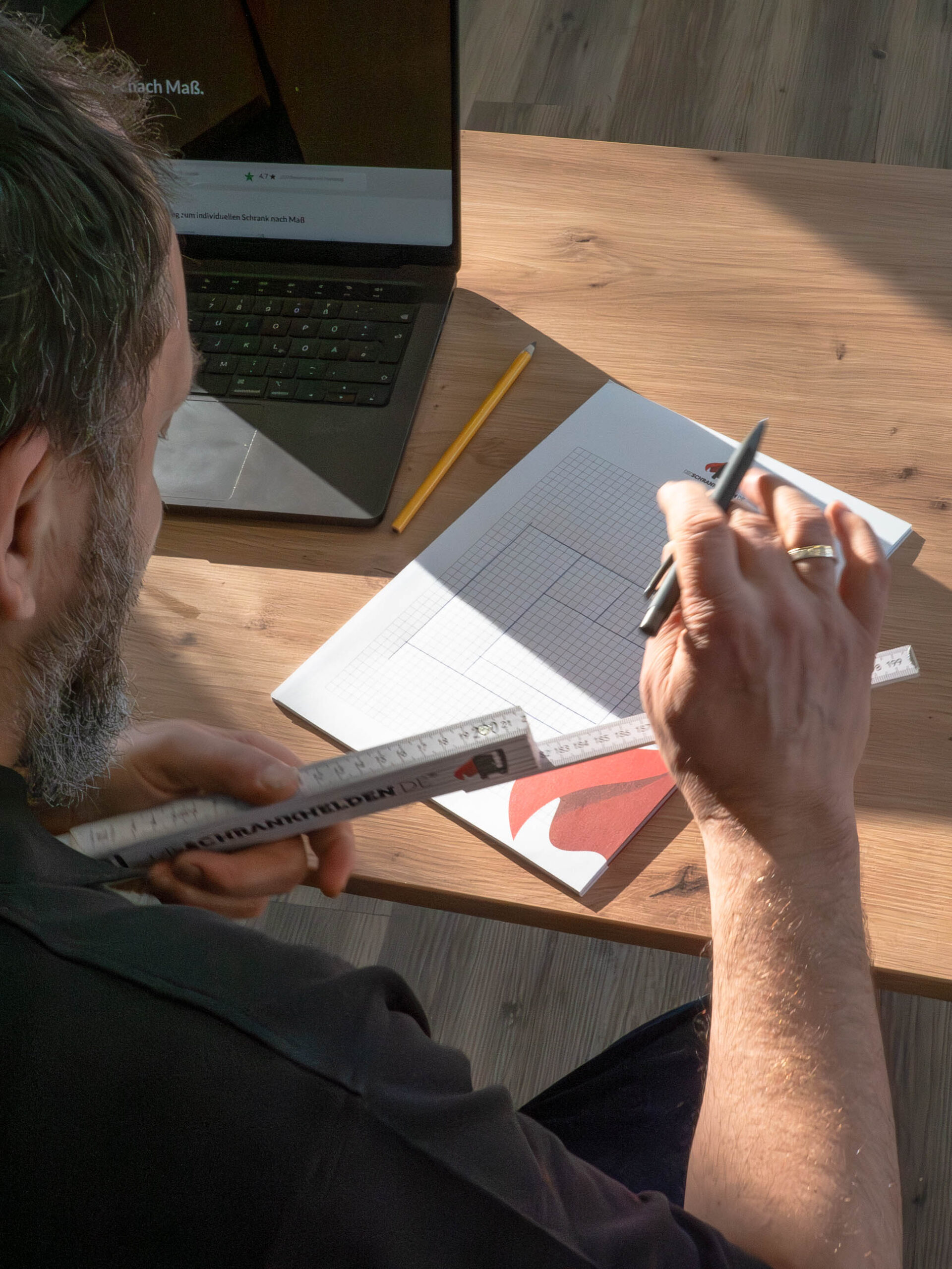 Tischler plant maßgefertigten Schrank mit Skizze, Maßband und Laptop am Tisch