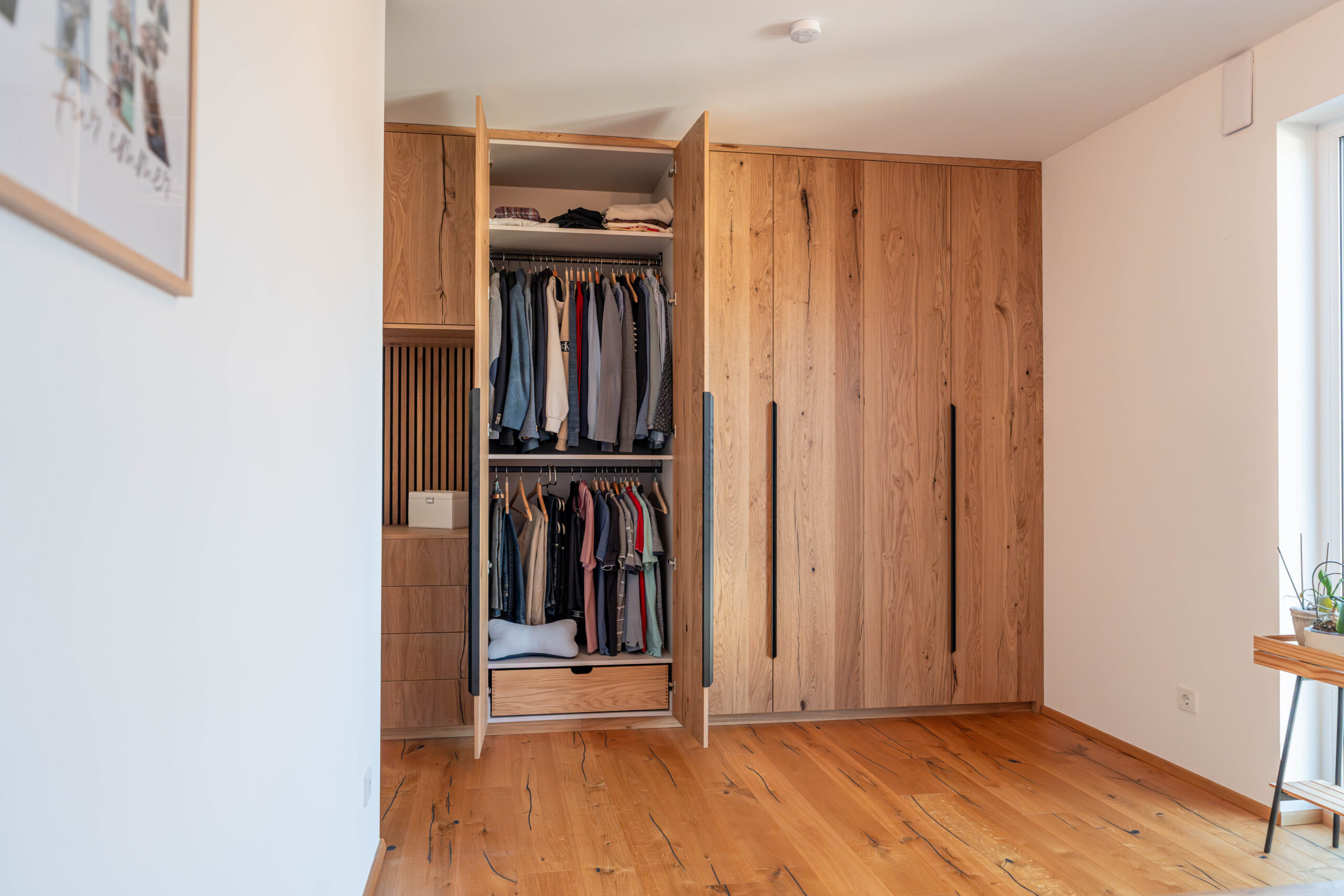 Geöffneter maßgefertigter Schlafzimmerschrank aus rustikaler Eiche mit zwei Kleiderstangen, Schubladen und schwarzen Griffleisten.